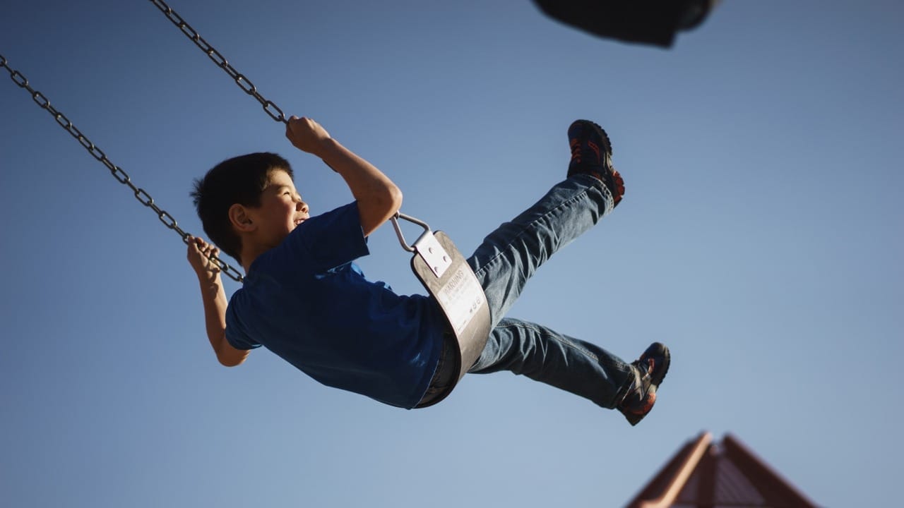 Kid swinging on a swing and smiling with a blue sky in the background