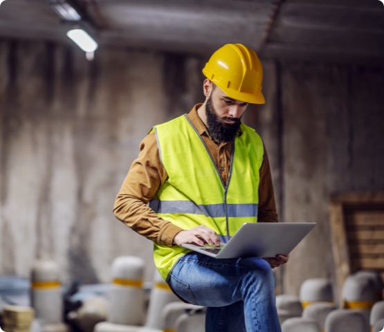 Construction supervisor in vest with helmet on head standing inside of underground parking lot in construction process and using laptop.