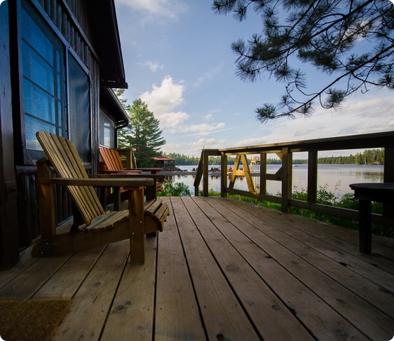 Two chairs on a wooden deck of a cottage facing a lake. In the background there’s a pier