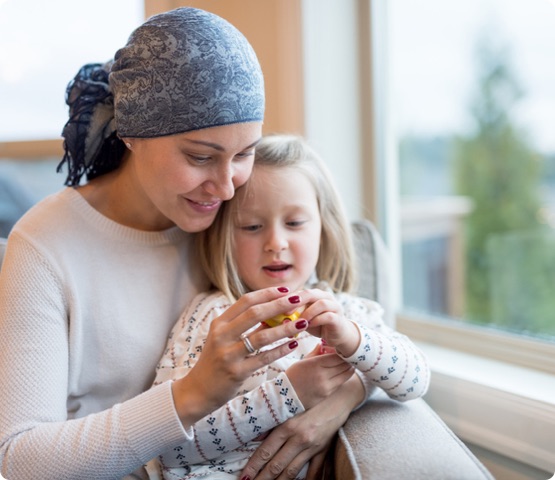 cancer patient holds daughter in her lap by their living window. They are holding and playing with a little plastic toy and mom is smiling. She is also wearing a headscarf.