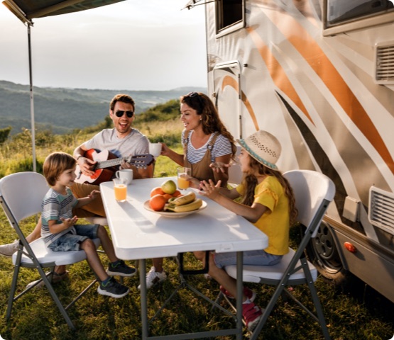 Family singing at picnic table by the camper trailer in nature. Man is playing a guitar.