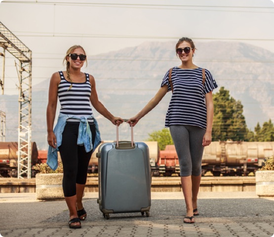Two people standing at the train station, holding onto luggage and smiling as a sign of excitement about the trip ahead