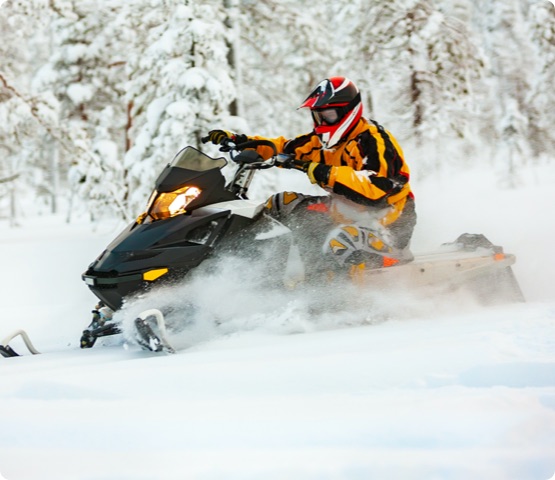 A person in the outfit of a racer in a yellow-black overalls and a red-black helmet, driving a snowmobile at high speed riding through deep snow against the background of a snowy forest.