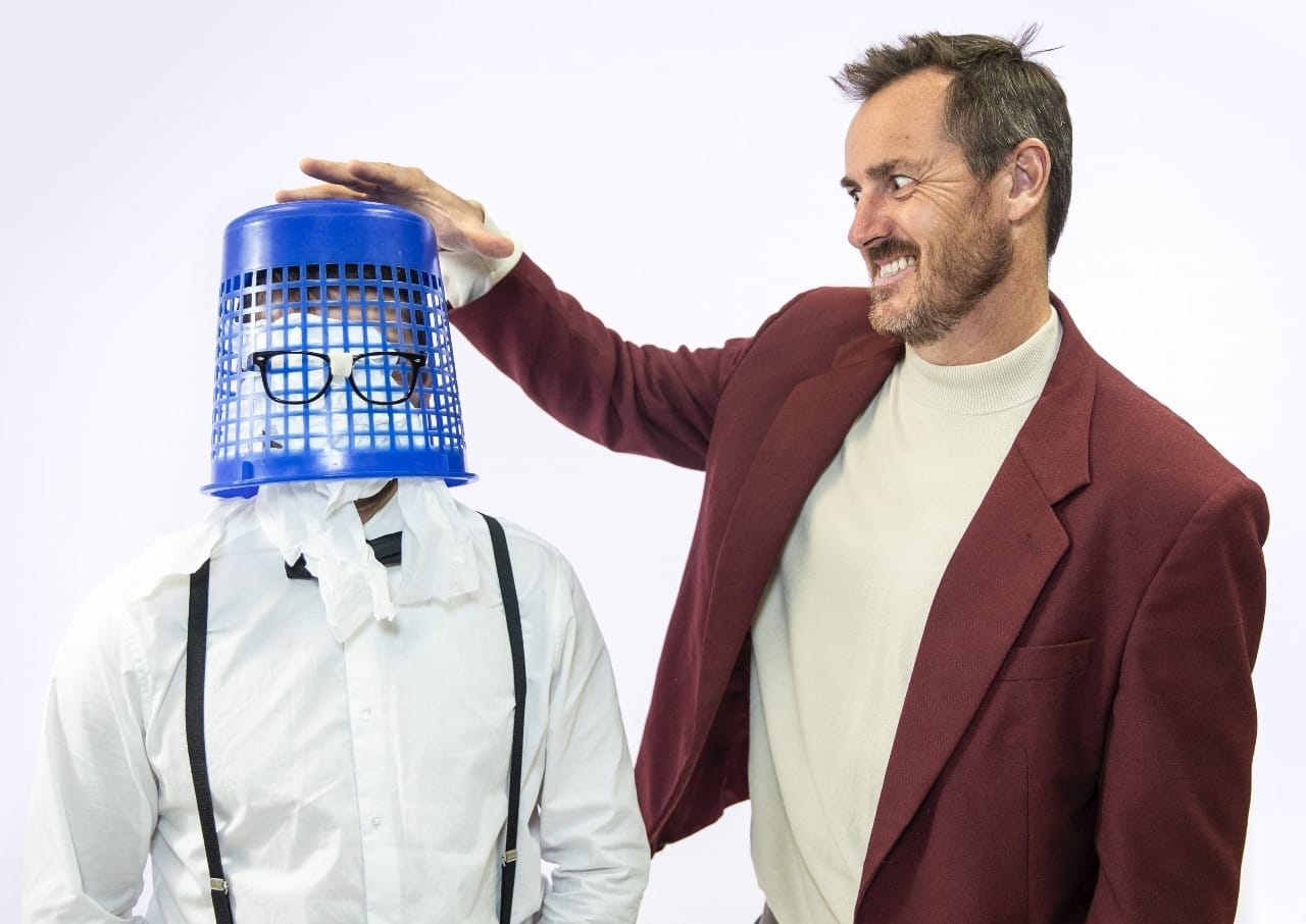 2 people goofing around. One person has waste basket on head with glasses stuck on the outside